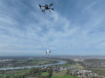 Drohne mit Messausrüstung über einer Flusslandschaft mit Feldern und Stadt im Hintergrund.